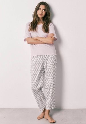 Ung kvinde står barfodet op ad en væg iført en lyserød top med korte ærmer og hvide løse bukser med et lille gråt mønster.