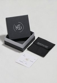 Fekete és szürke ékszerdoboz egy logóval, egy fekete tasakkal és egy kártyával, mindez egy fehér felületen fekszik.