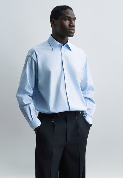 Homme portant une chemise boutonnée oversize bleu clair avec un pantalon noir plissé, mains dans les poches, regardant sur le côté sur un fond uni.
