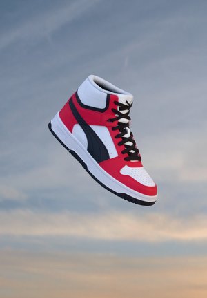 Basket montante blanche, rouge et noire avec des lacets noirs, présentée sur un fond de ciel nuageux.