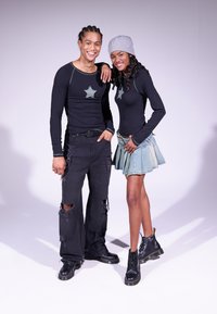 Două modele poartă bluze negre cu mânecă lungă, decorate cu patch-uri în formă de stea, jeans negri uzati și o fustă din denim. Femeia are o pălărie gri și cizme negre.