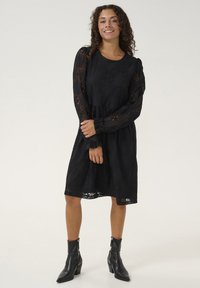 Čierne čipkované šaty s dlhými puff rukávmi, zúženým pásom a splývavou sukňou. Nosia sa s čiernymi členkovými topánkami s zipsovým uzáverom.