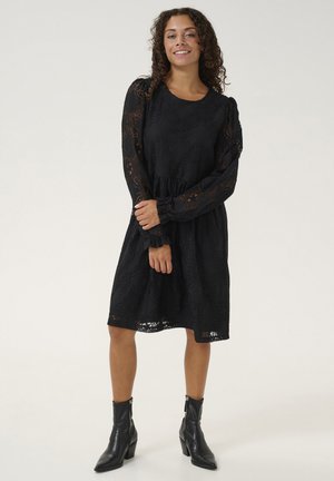 Zwarte kanten jurk met lange pofmouwen, een geplooide taille en een vloeiende rok. Draag het met zwarte enkelboots met een rits.