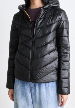 Femme portant une veste matelassée noire à capuche zippée, associée à un jean en denim bleu clair sur un fond uni.