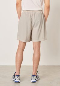 Osoba ubrana w beżowe prążkowane szorty, białą koszulkę i srebrno-niebieskie trampki, stojąca na jasnoszarej podłodze na tle jednolitej ściany.