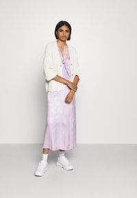 Crème gebreide cardigan, paars satijnen jurk met strikdetail en witte sneakers. De jurk heeft een subtiel patroon en een vloeiende uitstraling.