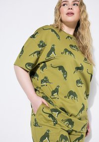 T-shirt in cotone verde con un design morbido, caratterizzato da un motivo ripetuto di leopardi in verde scuro e dettagli bianchi. Maniche corte, vestibilità rilassata.