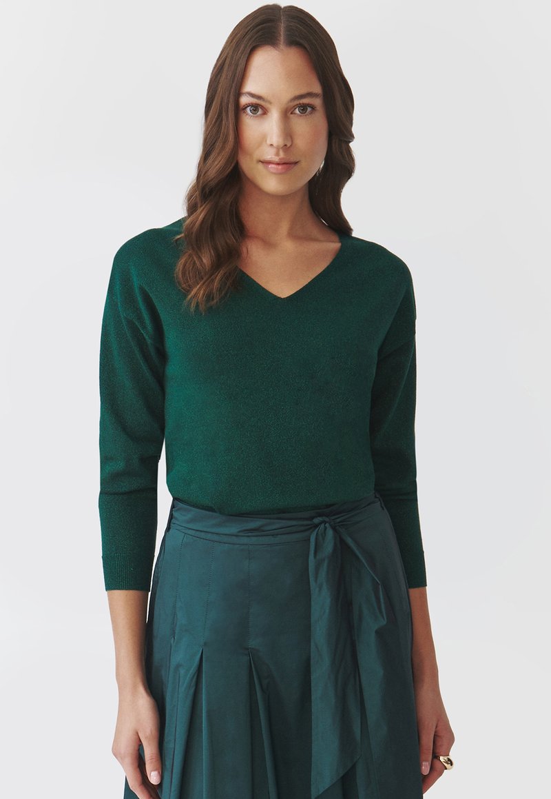 Groene, gestructureerde gebreide trui met een V-hals en lange mouwen, gecombineerd met een bijpassende teal rok met een strik om de taille en een geplooid ontwerp.