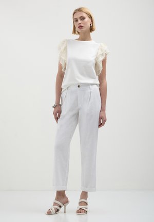 Donna in un completo tutto bianco composto da un top con maniche a sbuffo, pantaloni su misura, sandali con tacco e bracciale d'argento, davanti a uno sfondo bianco semplice.