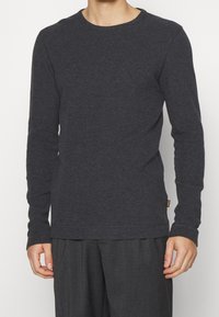 T-shirt à manches longues gris foncé avec un col rond. Fabriqué en tissu doux, présente des coutures visibles et une petite étiquette de logo à l'ourlet.