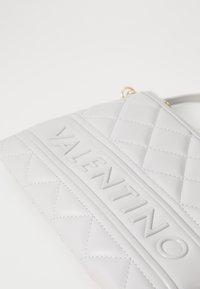 Saco de mão branco acolchoado com logotipo "VALENTINO" em relevo, detalhes em hardware dourado, textura em couro suave e um design estruturado.