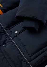 Navy sininen vuorattu takki, jossa on vetoketju, napit, ribbaatut hihansuut ja erottuva oranssi tikkaus tarratietuealueella. Tasainen pinta.