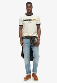 Camiseta crema con detalles en azul marino y logo, jeans azules, botas de cuero marrón y una camisa de franela a cuadros atada en la cintura.