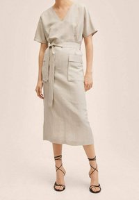 Robe en lin gris clair avec un col en V, des manches courtes, une taille ceinturée, et deux poches latérales. Associée à des talons noirs à lanières.