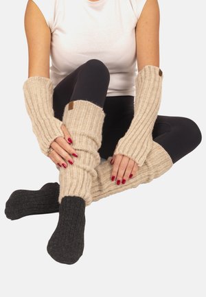 Frau sitzt im Schneidersitz und trägt beige gestrickte Beinwärmer mit dunkelgrauen Socken sowie passende beige fingerlose Handschuhe über schwarzen Leggings und weißem Oberteil.