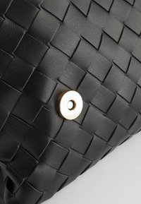Gros plan d’un motif tissé en cuir noir avec un fermoir magnétique rond doré sur un sac ou une pochette.