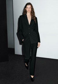 Black tailored suit featuring a deep V-neck blazer and loose-fit trousers. Smooth fabric with clean lines and minimal buttons for a sleek look.