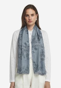 Ljusblå scarf med intrikata blommönster, fransade kanter och mjuk textur, bärs över en vit topp.