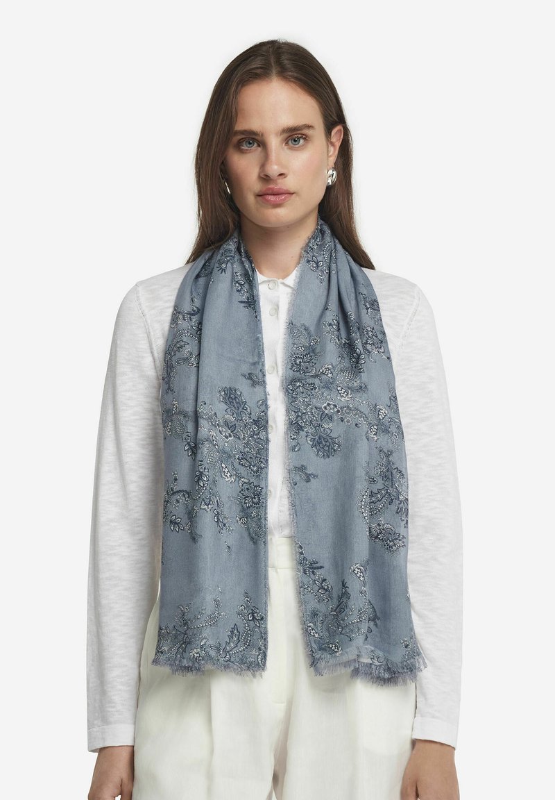 Ljusblå scarf med intrikata blommönster, fransade kanter och mjuk textur, bärs över en vit topp.