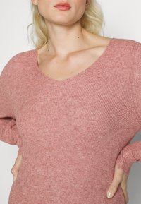 Blondynka w różowym swetrze z dekoltem w serek, z rękami na biodrach, na jednolitym tle.