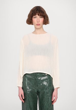 Cream-colored, sheer long-sleeve top with a wrinkled texture, worn over a dark gray tank top, paired with glossy green leather pants.