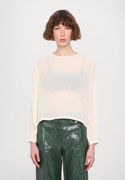 Haut long à manches transparentes de couleur crème avec une texture froissée, porté sur un débardeur gris foncé, associé à un pantalon en cuir verni vert.