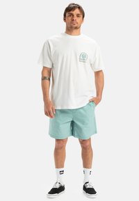 Valkoinen puuvillatee-shirt, jossa on pieni teal-välinen grafiikka, ja mintunväriset shortsit. Mustat lenkkarit valkoisilla pohjilla ja nilkkasukat.