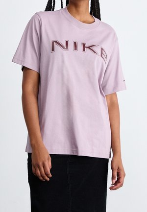 Osoba ubrana w jasnofioletową koszulkę z krótkim rękawem marki Nike z ciemnobrązowym logo oraz czarną spódnicę na jednolitym białym tle.