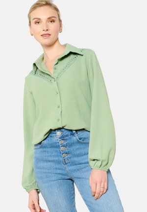 Camicia verde chiaro con colletto, caratterizzata da un bordo in pizzo decorativo sulle spalle. Maniche lunghe a palloncino e tessuto morbido. Indossata con jeans blu a vita alta.
