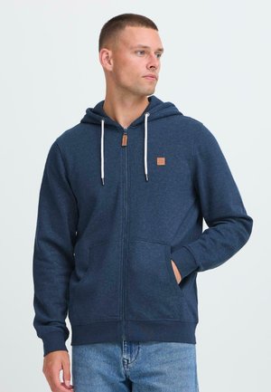 Jonge man in een marineblauwe zip-up hoodie met witte trekkoorden, staand met één hand in de zak, kijkend opzij.