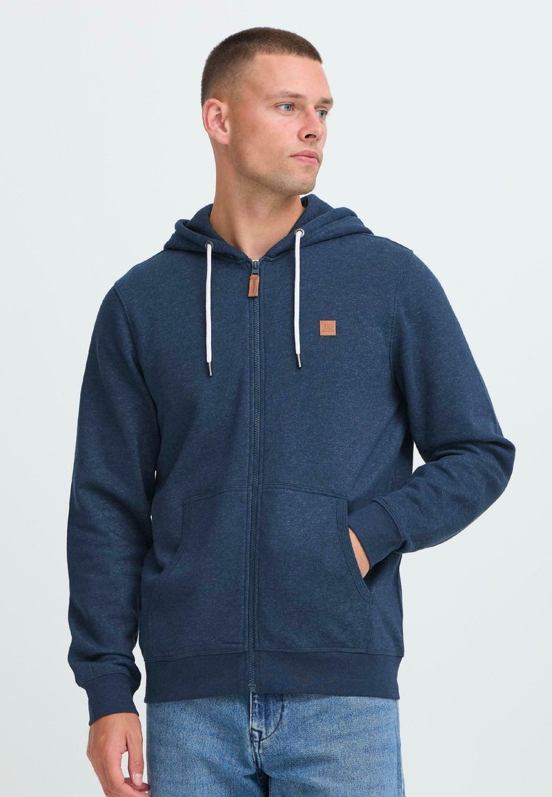 Mladý muž v námorníckom modrom zip-up mikine s bielymi šnúrkami, stojí s jednou rukou v vrecku a pozerá sa nabok.