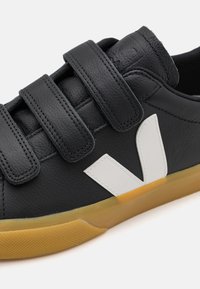Fekete bőr sneaker három állítható tépőzárral, fehér logóval és gumi talpval, amelynek színe barna.