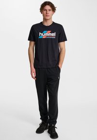 Czarna koszulka Hummel z kolorowym logo, w połączeniu z czarnymi spodniami dresowymi i czarnymi butami sportowymi. Prosty fason, luźne dopasowanie i gładka tekstura.