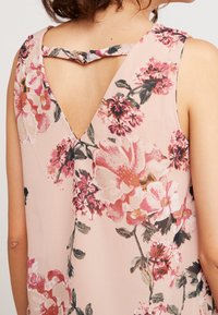 Vaaleanpunaisesta kankaasta valmistettu kukkakuvioinen hihaton pusero; siinä on V-muotoinen selkäosa ja kukkakuosi, jossa on vaaleanpunaisia, punaisia ja vihreitä yksityiskohtia.