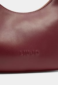 Borsa a mano in pelle bordeaux con una texture liscia e logo "LIU•JO" in rilievo. Design curvo con dettagli dorati lungo il bordo.