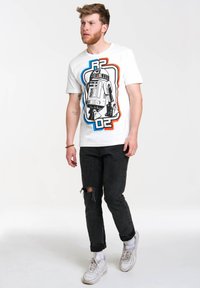 White t-shirt featuring a graphic of R2-D2 in black outlined design with blue and red accents. Worn with black distressed jeans and white sneakers.