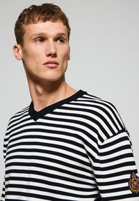 Striped sweater in navy and white with a V-neck, ribbed collar, and a patch accent on the left sleeve. Smooth, knit texture.