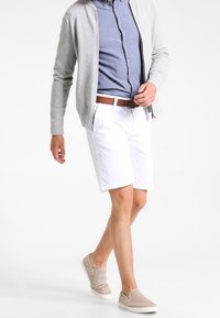 Lichtgrijze cardigan, blauw overhemd met knopen, witte korte broek en beige instapschoenen. De outfit heeft een bruine riem en een casual uitstraling.