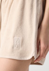 Bežīgas frotē šortas ar mīkstu tekstūru, kurām ir izšūts logo pie apakšmala un sānu šķiršanās, lai nodrošinātu ērtu kustību.
