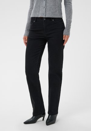 Kvinde iført sorte jeans med lige ben, grå knappet sweater og sorte spidse hælede støvler står mod en ensfarvet baggrund.