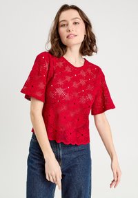 Haut en dentelle rouge tricoté avec des manches courtes, présentant des motifs floraux et un ourlet à volants. Assorti à un jean en denim foncé.
