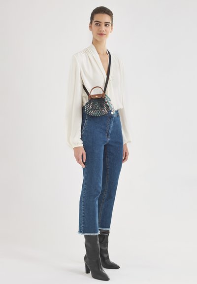 Black handbag with a mesh design, brown accents, worn crossbody; model in white blouse, high-waisted blue jeans, black heeled boots.