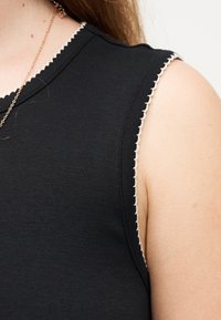 Κοντινό πλάνο στον ώμο ενός ατόμου που φοράει μαύρο αμάνικο τοπ με λευκή διακοσμητική κυματοειδή ραφή και χρυσή αλυσίδα λαιμού.