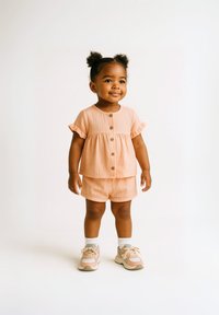 Ensemble en lin pêche comprenant un haut à volants et un pantalon court avec un bouton à l'avant. Associé à des baskets grises et roses et des chaussettes blanches. Texture lisse, coupe décontractée.