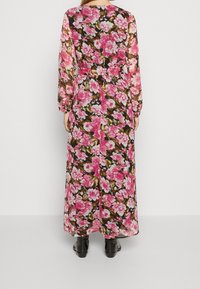 Kvinna klädd i en lång, genomskinlig blommig klänning med rosa blommor och gröna blad, i kombination med svarta ankelstövlar, som står mot en enhetlig vägg.