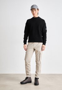 Suéter de punto negro, pantalones cargo beige con bolsillos laterales y gorra gris. El calzado son botas oscuras y resistentes con cordones y soporte para el tobillo.