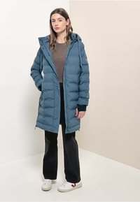 Abrigo puff de color teal con capucha, que presenta un cierre completo, bolsillos laterales y puños acanalados. Se lleva sobre un suéter marrón y pantalones negros.