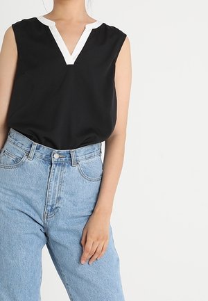 Kobieta w czarnej bluzce bez rękawów z białym wykończeniem w kształcie litery V na dekolcie oraz w jasnoniebieskich dżinsach z wysokim stanem, z ręką spoczywającą na udzie na białym tle.