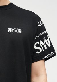 Μαύρο κοντομάνικο μπλουζάκι με λευκό λογότυπο "Versace Jeans Couture" στο στήθος και επαναλαμβανόμενο κείμενο λογότυπου στο μανίκι.