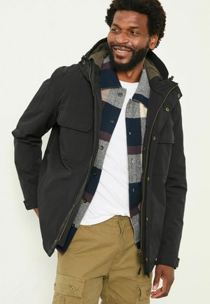 Black jacket with a hood, featuring front pockets and a zipper. Worn over a gray and blue plaid shirt, paired with beige cargo pants.